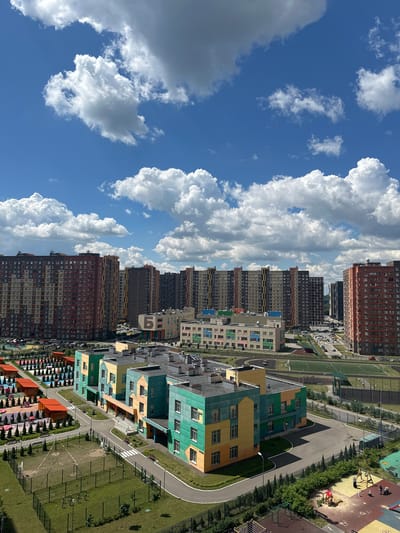 The image is an aerial view of a modern residential complex in Mirny, Russia. The scene is dominated by tall, colorful apartment buildings with a variety of facades, including brick, dark grey, and brightly colored panels in green, yellow, and orange. These buildings are arranged in rows, with some featuring balconies and numerous windows.
Between the buildings, there are paved pathways, green spaces with trees and grass, and a playground with colorful equipment and visible benches. A sports field, possibly a soccer pitch, is also visible. The architecture suggests a relatively new development.
The sky is a vibrant blue, dotted with large, fluffy white clouds, indicating a clear and pleasant day. The sunlight casts distinct shadows on the ground, suggesting it is daytime, likely late morning or early afternoon.
No people are clearly discernible in the image, but the presence of playgrounds and apartments implies a residential area. The overall impression is one of a well-planned, modern urban environment set against a beautiful sky. There is a partially visible number "5" on one of the buildings.