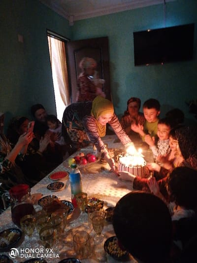 An indoor scene in what appears to be a residence in Samarkand, Uzbekistan, depicts a gathering of approximately twelve individuals, predominantly women and children, around a table. The walls are a muted teal color, with a dark wooden door partially open to the left revealing a light-colored curtain. A flat-screen television is mounted on the wall above the gathering.
The central focus is a decorated, round cake with multiple lit candles, positioned on a white patterned tablecloth. A woman wearing a yellow hijab and a patterned long-sleeved tunic is leaning over the cake, her hands near the candles, suggesting an act of lighting or presentation. She is smiling towards the right side of the frame.
Several children, including a young boy in a light green shirt, are gathered closely around the cake, looking at the lit candles with expressions of excitement. Another young child in a patterned top is also visible next to the boy. On the left, a woman is holding a smartphone, directed towards the cake, indicating she is recording or photographing the event. In the background, a blurred adult figure is visible near the door, and another adult holds a small child.
The table is set with various items: a dark red liquid in a glass pitcher, multiple clear glasses (some containing liquid), small bowls of snacks such as nuts or dried fruit, fresh fruits including red apples and pomegranates, and a plastic bottle containing a clear beverage. The primary light source appears to be the candles on the cake, casting a warm glow on the immediate area and faces.