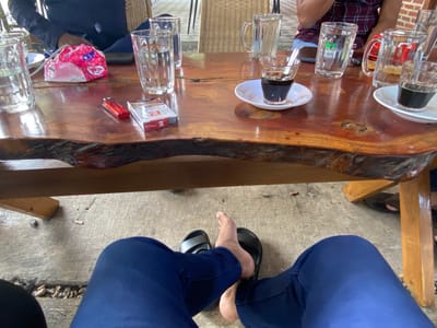 This image depicts a scene at a table, likely outdoors, in Aluepiet, Indonesia. The focus is on a wooden table with various items on it and the lower body of a person wearing blue pants and black sandals.
On the table, there are several clear glasses, some filled with water, others with dark liquid appearing to be coffee. A small white saucer holds a glass of coffee with a spoon. A pack of cigarettes and a red lighter are also present. A pink and white patterned wrapper, possibly for tissues or snacks, is near the glasses. In the background, parts of people can be seen, including a person wearing a dark shirt and another wearing a maroon and white checkered shirt. The table appears to be set up in a casual outdoor area, possibly a cafe or roadside eatery, with concrete flooring visible below. There are also some discarded cigarette butts on the concrete. The lighting suggests it is daytime.