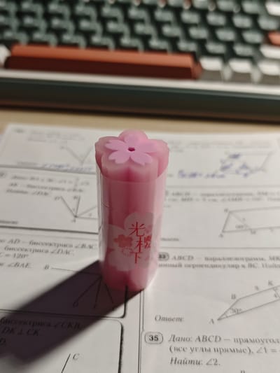 The image shows a close-up, eye-level shot of a pink, cherry blossom-shaped eraser resting on a sheet of paper with mathematical diagrams and text. The eraser is positioned vertically and slightly off-center to the right. The paper appears to be a worksheet or textbook page with geometry problems, indicated by the presence of lines, angles, and mathematical notation in Cyrillic. A blurry keyboard, likely a computer keyboard, is visible in the background, suggesting an indoor setting, possibly a desk. The lighting in the image creates a shadow from the eraser cast onto the paper. The image does not contain any identifiable landmarks or features that would allow for its location to be determined in Svietlahorsk, Belarus, or any other specific city.