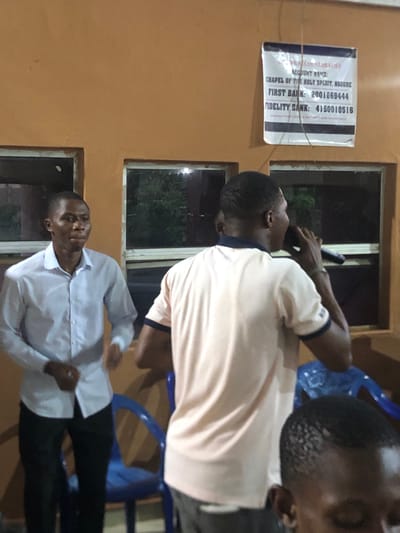 The scene appears to be indoors, potentially a community center or a religious building in Nsugbe, Nigeria. The walls are orange-toned. Two men are visible; one is wearing a white shirt and black pants, while the other is wearing a light peach shirt with a dark collar. A poster on the wall reads "ACCOUNT DETAILS, ACCOUNT NAME: CHAPEL OF THE HOLY SPIRIT, NSUGBE, FIRST BANK: 2001669444, FIDELITY BANK: 4150010516". There are two windows present. One of the men appears to be speaking into a microphone. Blue plastic chairs are also visible.