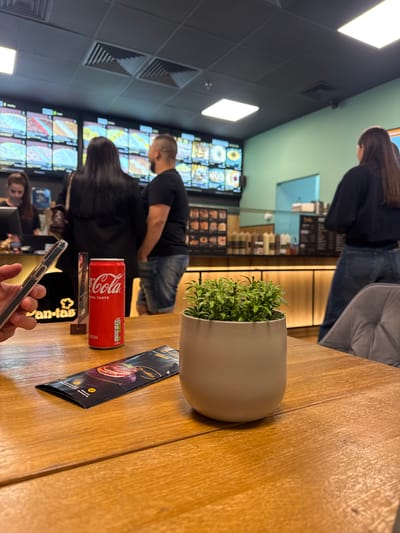 The image shows a scene inside a restaurant, possibly in Yambol, Bulgaria, given the "Pan-tas" menu on the table. A wooden table takes up much of the foreground, on which sits a potted plant, a Coca-Cola can, and a black menu. A person's hand is holding a smartphone. In the background, people are standing in line at the restaurant's counter, in front of a menu board with food pictures. The restaurant has a modern interior with grey walls, recessed lighting, and a green accent wall.