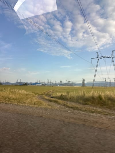 The image displays a landscape with a clear sky filled with scattered clouds. Tall power lines and pylons stretch across the frame, indicating an area with significant electrical infrastructure. The foreground on the right shows a dirt or gravel road, with dry grass and vegetation along its edge, suggesting a rural or undeveloped setting. In the distance, more power lines and structures are visible, along with rolling hills or mountains under a hazy sky. The overall scene conveys a sense of open space and industrial presence within a natural environment. The lighting suggests it is daytime, likely late morning or early afternoon given the bright sky. There is no visible text in the image.