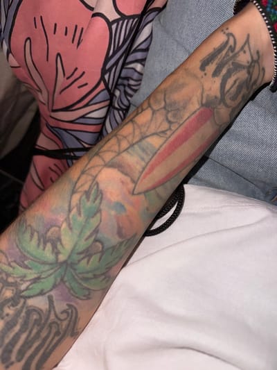 A close-up shot features a person's tattooed forearm, angled diagonally across the frame. The arm is extensively covered with multiple tattoo designs. On the upper part of the forearm, near the wrist, are dark grey, script-like letters that are difficult to definitively decipher, possibly forming "NO" or "NCS." Below these letters, a distinctive red and white striped element, resembling a knife blade or a stylized flag, is visible, surrounded by a faint, web-like or scaled pattern. Further down the arm, a prominent green leafy design with watercolor-like splashes of purple and blue hues is present. Towards the lower portion of the forearm, darker, more abstract tribal-style patterns or wings can be seen.
The arm is set against a backdrop of various fabrics: a textured blue denim material (likely a jacket) is visible on the upper left, and a plain white t-shirt or garment occupies the right side of the frame. A black braided cord is nestled between the arm and the white fabric. In the lower left, a soft pink and purple fabric with a stylized floral or abstract pattern is visible, possibly part of another piece of clothing or a blanket. The image is a close-up, offering no clear indication of a specific activity or the time of day.