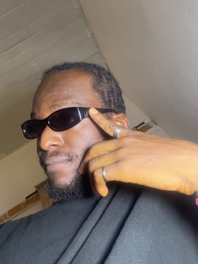 The image shows a close-up, low-angle shot of a Black man wearing sunglasses and a black garment. He has braids and a beard, and is wearing two silver rings on his right hand. His hand is positioned near his face, obscuring part of it. The background appears to be an indoor setting with cream-colored walls and wooden paneling. The lighting suggests it might be daytime, but the interior setting makes it difficult to determine the time of day. There are no obvious location cues to suggest it is specifically Ban, Nigeria, beyond the provided context. The man's expression is not clearly visible due to the sunglasses and hand. The overall mood is somewhat enigmatic.