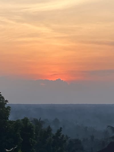 The image depicts a panoramic natural landscape in Nguzu Edda, Nigeria, captured during either sunrise or sunset. The upper third of the frame is dominated by a sky that gradients from a pale, warm yellow-orange at the top to a rich, intense orange-red at the mid-level. A prominent, dark horizontal band of dense cumulus-like clouds extends across the middle of the frame, partially obscuring the sun, which appears as a bright, glowing orange-red source just above the cloud line in the center.
Below this cloud layer, the atmosphere appears hazy, rendering the distant landscape in successive layers of muted blue-gray tones. This midground comprises a vast expanse of rolling terrain covered in dense, indistinct vegetation, likely forests or agricultural areas, stretching towards the horizon. No specific structures or land features are clearly discernible in this hazy distant view.
The foreground, primarily on the left and bottom sections, features the dark silhouettes of dense tree foliage, indicative of vegetation close to the camera. The leaves are undifferentiated due to the backlighting conditions. A dark, indeterminate mass, possibly a structure or additional dense foliage, is present in the lower right corner. No people, animals, or distinct man-made objects or structures are visible within the frame. The overall lighting conditions suggest a twilight period, characterized by soft, warm light from the low-lying sun and significant atmospheric haze.