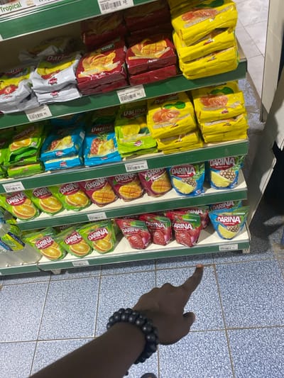 The image displays an indoor scene, likely within a supermarket or convenience store in Goderich, Sierra Leone. The primary focus is on a well-stocked display of instant drink mixes arranged on multiple green and white shelves, with a person's arm and hand visible on the right side of the frame.
The shelves are filled with numerous packets of instant drink mixes, predominantly branded "DARINA". Various flavors are discernible by their packaging colors and labels: Mango (in dark red packets), Pineapple (in blue packets), Strawberry (in red packets), Orange (in yellow and orange packets), Lemon (in light green packets), and Tropical (in white packets). Some packets also explicitly mention "Instant drink" or "Aloevine". Price tags attached to the shelves show visible barcodes and numerical values such as "93", "9.0", "50", "4", and "30".
On the right side, a dark-skinned arm and hand are seen, adorned with a dark, multi-beaded bracelet on the wrist. The hand is pointing to the right, suggesting the person is indicating an item or a direction. The floor is tiled with a pattern of light blue and white squares. The overall lighting suggests an interior setting with artificial illumination.