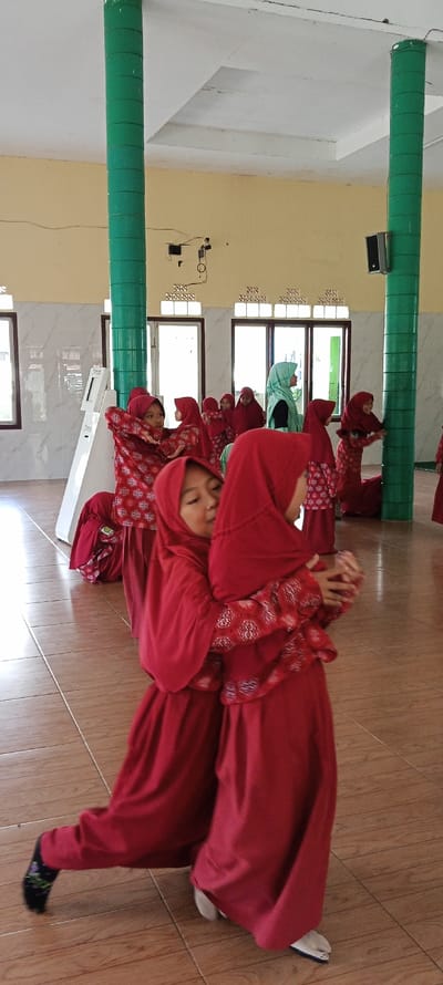 The image shows a group of young girls in red and white clothing, standing in a room with tiled floors and a white ceiling. Two girls are at the center of the image, with one in a bright red hijab and the other wearing a patterned red and white hijab. They are embracing each other, with one's arm around the other's shoulder. The girls in the background are also in similar red and white clothing, with some wearing green hijabs. There is a white and green pillar in the middle of the room, and windows are seen in the background. The coordinates provided are for the town of Cangkuang, Indonesia, where this image was taken. The scene is casual, likely captured during a moment of playtime or a break from school. There are no signs of forced interaction or discomfort.