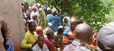 A group of people gathers outdoors, primarily Black individuals of varying ages, dressed in casual clothing and colorful, patterned fabrics. Some wear head coverings, like turbans and caps. The crowd is dense, with some individuals seated and others standing. To the left, a light brown wall is visible, and green trees fill the background. The scene seems to capture a community event or gathering.