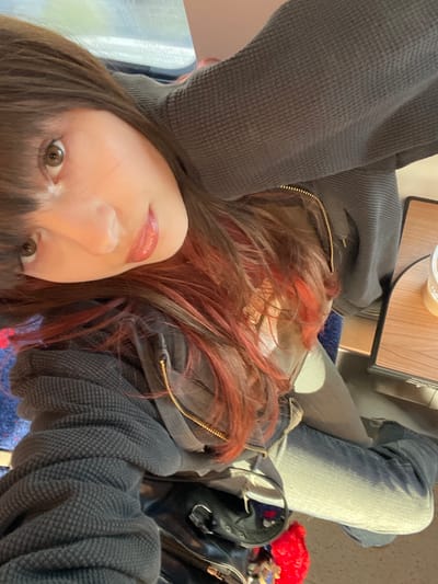 A close-up selfie shows a person with light skin, brown hair with red highlights, and visible makeup on their lips. They are wearing a gray waffle-knit zip-up jacket and blue jeans. In the background, there is a small wooden table with a drink on it and a glimpse of the train or bus seat. The presence of Hebrew characters on the table suggests that the location is likely in Israel, and as there is no explicit data linking the image to Ramat HaSharon, it is not possible to confirm the location. The subject appears to be seated on public transportation. A purse and a red bouquet are partially visible near the subject.