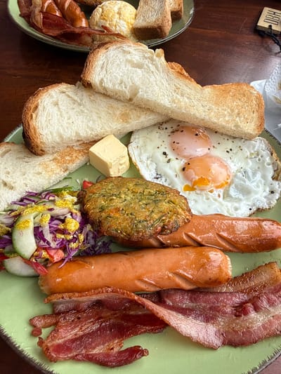 A hearty breakfast plate showcases toasted bread slices, two sunny-side-up eggs, two cooked sausages, and crispy bacon strips. A green fritter and a cube of butter add to the ensemble. Garnishing the plate is a small salad of red cabbage and cucumber with a yellow dressing. A side plate offers more bread and bacon. The meal sits on a wooden surface, suggesting a casual dining setting.