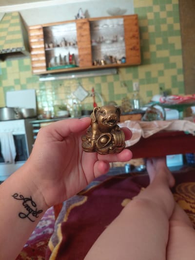 The image shows a close-up of a person's hand holding a small, ornate brass figurine. The figurine appears to be a stylized animal, possibly a cat or a dog, with large eyes and a textured body. The person's wrist is also visible, with a tattoo that reads "Angel" in script. The background is a tiled floor with a pattern of green and yellow squares, and some wooden shelves filled with small items. There is also a glimpse of a person's legs in the upper right of the frame, suggesting a seated or reclining position. The lighting suggests it is daytime. The setting appears to be indoors, likely a domestic environment in Marynivka, Ukraine. The image is taken from a slightly overhead angle.