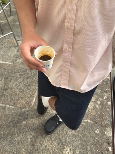The media file shows a person sitting outdoors, holding a small white paper cup filled with a dark liquid, likely coffee. The person is wearing a light pink checkered button-down shirt and dark blue jeans, with white socks and black slip-on shoes. Their left arm and hand are visible, with the hand gently holding the cup. The background is a rough, greyish stone surface, possibly a sidewalk or plaza, with a metallic railing visible at the bottom left.
The lighting suggests it is daytime, with sunlight casting shadows. The overall scene appears casual and relaxed, with the person taking a break to enjoy a drink. The text "CAFE" is visible on the side of the paper cup. Given the location context of Santiago de Compostela, Spain, this could be a tourist or pilgrim taking a moment to rest during their travels. There are no explicit cues about the weather, but the bright lighting suggests it is likely sunny. The emotion conveyed is one of calm and perhaps slight weariness, common for those undertaking long journeys.