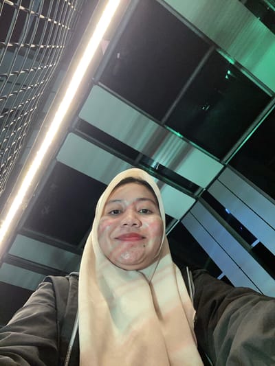This media file shows a selfie of a person in Banjarbaru, Indonesia. The person is wearing a light-colored hijab and a dark jacket. Their face is partially visible, with a playful expression. The background features an abstract architectural design with a combination of dark and light-colored panels, possibly part of a building or an outdoor structure. A long, bright light fixture is visible in the lower part of the frame, illuminating a metal grate or railing in front of it. The lighting and overall composition suggest it might be evening or nighttime. There is no visible text in the image.