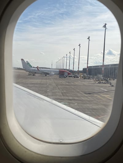 The image shows an airplane parked at an airport, viewed through an airplane window. The airplane is white with red and green accents. In the background, there is a long building with a red and white striped facade, and a series of tall poles with lights on them. The ground is paved and marked with yellow lines. The sky is blue with some clouds. The wing of the airplane from which the photo is taken is visible in the foreground, also white. The scene is likely at Berlin Brandenburg Airport (BER) in Schönefeld, Germany, given its recent opening and location. There are no people clearly visible in the image, but there are some service vehicles and equipment around the parked airplane. The overall impression is a view of an airport tarmac under a partly cloudy sky.
