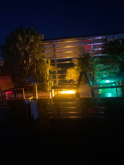 This image depicts an outdoor seating area at night, illuminated by warm yellow and cool green lights. In the foreground, dark steps lead up to a paved area with furniture. A wooden chair with a slatted seat and back is visible on the left, next to a round glass-topped table with a metallic base. Behind the table, a potted snake plant with distinctive sword-like leaves stands. Further back, a tall, dense evergreen tree is partially lit, casting shadows.

Above the seating area, a structure with horizontal beams is visible, illuminated by rows of orange and pink lights. Several tropical plants with frond-like leaves are placed in this upper level, bathed in green light. The overall ambiance suggests a bar, restaurant, or lounge setting. The sky is a deep, dark blue, indicating nighttime. The image does not contain any people or explicit actions. The scene is clean and appears well-maintained, with no visible signs of neglect or disarray. The location in Sabon Gari West, Nigeria, suggests a possible urban or suburban environment.