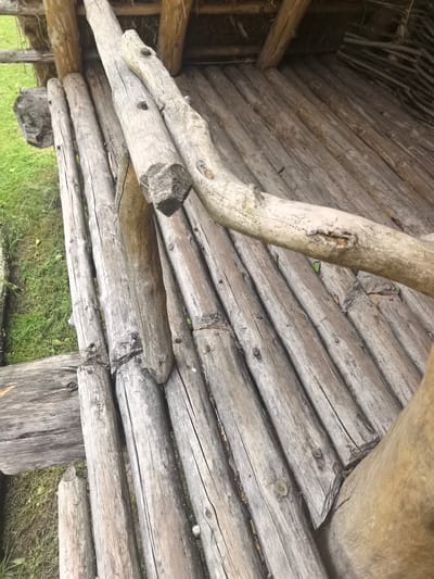 This is a close-up, eye-level, full shot of a rustic wooden structure, likely part of a historical reenactment or a traditional building in the Камянюцкі сельскі Савет, Belarus. The dominant feature is the rough-hewn, weathered wood, arranged in a diagonal pattern across the frame. Several logs of varying thicknesses form the surface of the structure, their grey, aged appearance marked by visible grain, cracks, and knots.
In the foreground, a thick, lighter-colored log runs diagonally from the upper right to the lower left, obscuring some of the underlying structure. There are no people visible in the image, and no actions are depicted. The environment surrounding the structure is suggested by the presence of green grass and a hint of what appears to be straw or thatch roofing in the background. The lighting is natural, casting subtle shadows that emphasize the texture of the wood. The overall impression is one of age and natural materials, evoking a sense of history and a connection to traditional construction methods.