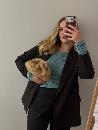 A woman is taking a selfie in a mirror. She is wearing a dark pinstripe blazer over a teal ruched top and black pants. She is holding a tan knitted clutch bag. Her hair is blonde and wavy, and she is wearing light makeup. The background is a plain light-colored wall. The location is Dreiliņi, Latvia.