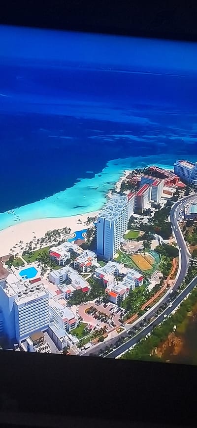 The image is an aerial view of a coastal resort area. A vast expanse of deep blue ocean dominates the top left of the frame, transitioning to a vibrant turquoise shallow water area along a sandy beach. Numerous resort buildings, primarily white and light-colored high-rises and lower-rise complexes with red accents, are situated along the coastline and inland. Lush green vegetation and palm trees are interspersed among the buildings. A winding road with a few cars is visible, bordering the resort complex. Several swimming pools, including one with a unique kidney shape, are present within the resort grounds. There are also tennis courts and what appears to be a parking lot filled with vehicles. The scene depicts a developed tourist destination with extensive beachfront properties and amenities. The location is not in Bila Tserkva, Ukraine, as that city is landlocked and does not have a coastline.