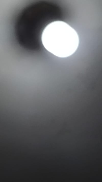 The image is a close-up, out-of-focus shot of a ceiling fan light. A bright white lightbulb is visible, partially obscured by a dark shadow that appears to be a portion of the fan's housing or blades. The background is a blurred gradient of gray, suggesting the ceiling. There are no people, animals, or other objects visible in the frame. The image lacks any identifiable landmarks or details that would allow for its specific location to be determined within Lubuk Pakam, Indonesia, or any other city.
