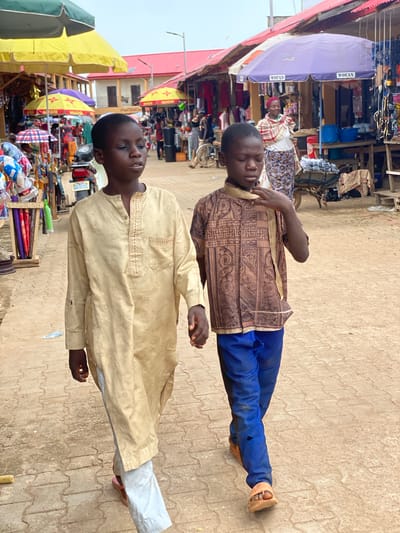 The image shows two young boys walking on a paved pathway in an outdoor market. The boy on the left is wearing a long, light-colored tunic and light-colored trousers. The boy on the right is wearing a patterned brown shirt and blue trousers. They are walking towards the viewer, slightly angled to the left. In the background, there are market stalls with awnings, some displaying merchandise such as clothing and household items. A woman in traditional attire is visible behind a purple awning. The ground is paved with interlocking bricks. The location is identified as a market in Kafanchan B, Nigeria.