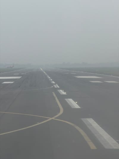 The image displays an airport runway or taxiway extending into the distance under conditions of extremely heavy fog or haze, significantly reducing visibility. The perspective is from an elevated vantage point, looking down the length of the paved surface.
The dark grey asphalt is clearly marked with various white rectangular patterns and a system of yellow lines. In the foreground on the right, a prominent wide, curved yellow line is visible, while parallel straight yellow lines delineate the left edge of what appears to be the main pathway. Embedded along these markings and edges are numerous small, bright yellow and white runway lights, illuminating the immediate foreground before their light dissipates into the dense atmospheric obscurity.
The heavy fog or haze is the most dominant feature, obscuring the background entirely and creating a uniform light grey sky that merges seamlessly with the misty horizon. Far in the distance, on the left side of the runway, a faint, small reddish-orange object, possibly an aircraft or a ground vehicle, can be vaguely discerned, almost completely swallowed by the low visibility.
The scene captures an active airport environment operating under challenging weather conditions, likely during dusk, dawn, or night, or a severely overcast day, highlighting the reliance on artificial lighting for navigation. No discernible text is visible in the image. The setting is an airport in Pajang, Indonesia.