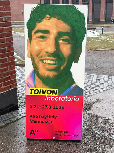 A tall, vertical advertising sign is positioned on a wet, paved surface next to a red-brick building. The sign features a large portrait of a smiling male with fair skin, dark eyes, and dense, curly green hair. He has light stubble and wears a collared garment with iridescent blue and purple tones. Below the portrait, a bright red-to-magenta gradient background displays white and yellow text. The yellow text reads "TOIVON", followed by white text "laboratorio". Further white text states "5.2. - 27.3.2026", "Koe näyttely Marsiossa." (Experience the exhibition at Marsio.), and an "A''" logo, along with the URL "aalto.fi/fi/toivonlaboratorio". The presence of "Marsio" and the Aalto University domain indicates the location is the Aalto University Otaniemi campus in Espoo, Finland. The ground around the sign is a mix of light-colored patterned pavers and dark asphalt, scattered with small dark pebbles, appearing wet. In the background, beyond a thin strip of snow-dusted ground and dark bollards, stands a large, dark, multi-story modern building. Two indistinct human figures walk in the far background. The sky is overcast, providing diffused lighting.