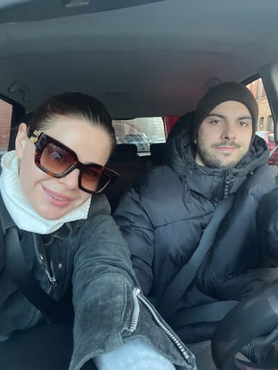 Two people pose inside a car, likely taking a selfie. The woman in the foreground wears large, tortoise-shell sunglasses, a white turtleneck sweater, and a gray jacket. A safety belt is visible across her torso. Behind her, a man wears a black beanie and a puffy black coat with a seatbelt crossing his chest. The interior of the car is gray, and a blurry exterior view, possibly a street scene, can be seen through the windows.