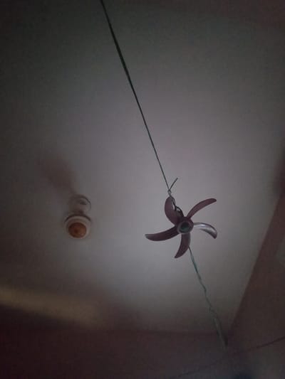 In a dimly lit room, a ceiling fan with five dark purple blades is suspended by a green string. The fan is positioned in the foreground, angled downwards. In the background, another ceiling fixture, possibly a light or a smaller fan, is visible. The ceiling is a pale color, and shadows are cast across it. The scene is indoors, in a residential setting. The location is identified as Peshawar City Tehsil, Pakistan. There are no people or animals visible in the image.