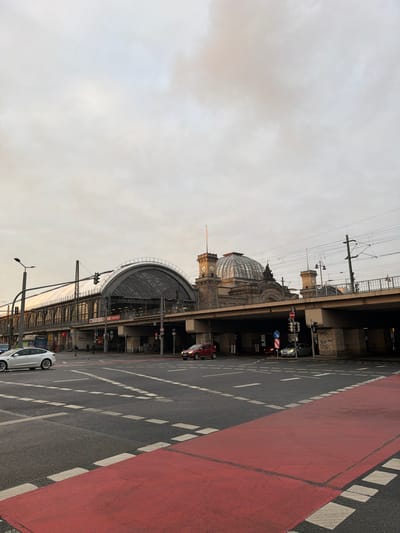 The image displays an urban street intersection in Dresden, Germany, with the Dresden Hauptbahnhof (main train station) prominently in the background.
The foreground consists of an asphalt road marked with white dashed lines, indicating lanes and turn designations. A distinct red-painted lane or pathway, likely for bicycles, spans the immediate bottom of the frame. On the road, a white Tesla Model S is visible on the far left, oriented to proceed left. A red SUV is positioned further right in the mid-ground, facing right, and a dark-colored sedan is to its right, also facing right; both appear stationary or moving slowly at the intersection. Traffic lights and street lamp poles are positioned at the intersection.
The Dresden Hauptbahnhof dominates the background as an elevated structure. The station's left section features a large, arched roof constructed of glass and metal, covering railway tracks. To the right of this arch, a prominent, textured dome is visible, flanked by two square, stone clock towers. The tower on the viewer's right displays a visible clock face. The station's foundation consists of concrete and stone supports, forming underpasses beneath the elevated tracks. Several individuals are discernible walking under the elevated structure on the left side. Overhead power lines and associated utility poles are present above the street level. The sky is uniformly overcast, appearing light grey and white.