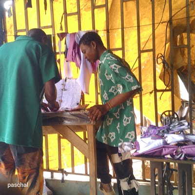 Two men are working in a small shop in Nsugpe, Nigeria. One man, seen from the back, wears a green shirt and printed pants, is bent over a wooden table, examining or working on a flat object. The second man, to the right, also stands at the table. He wears a green shirt with a pattern and dark pants. He has a bracelet on his right wrist. Behind them, a yellow plastic tarp is stretched across a metal grid. Clothes hang to dry. A sewing machine and folded fabrics rest on a table.