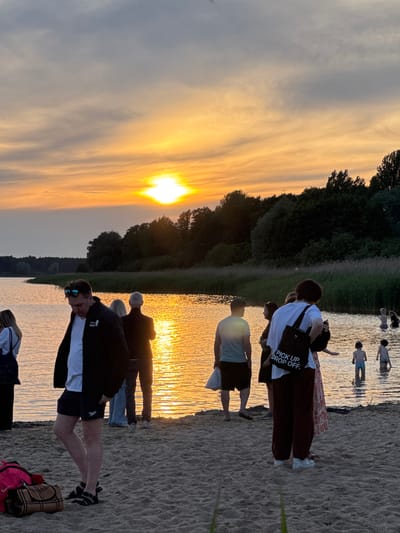 The image depicts a group of people on a sandy beach next to a body of water at sunset. The sky is filled with dramatic clouds illuminated by the setting sun, casting a golden and orange glow on the water. In the foreground, several individuals are standing on the sand, some appearing to be conversing or observing the sunset. Further back, a few people are wading in the shallow water. The background shows a line of trees and reeds along the water's edge. The overall atmosphere is serene and evokes a sense of a relaxed evening. This scene is likely located at a beach in the city of Tallinn, Estonia, given the commonality of such picturesque sunset views over coastal areas or lakes within or near the city.