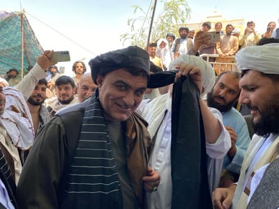 A group of men stand together in a crowd. The main subject is a man in a black turban and striped black and white scarf. He is smiling and adjusting his turban. He is surrounded by other men in traditional clothing. One man in a white turban is holding a black scarf and looks at the main subject. Another man to the right of the main subject is holding a white phone and talking. In the background, a few people are on a balcony. The image was likely taken in Zaker-E Sharif, Afghanistan, at the coordinates 31.547, 65.761. The scene appears to be a public event, possibly a religious ceremony.