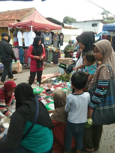 This image depicts a bustling outdoor market scene in Baleendah, Indonesia, under what appears to be an overcast or cloudy sky. Numerous individuals, predominantly women and children, are engaged in various market activities, suggesting a local community market.

In the mid-ground, a prominent red canopy stall features a variety of clothing items, including white t-shirts, striped shirts, and graphic tees, displayed on racks. A woman in a red long-sleeved tunic and black hijab stands in front of this stall, appearing to be a vendor. To her left, a man in a black jacket and cap walks past. Further right, another woman in a black hijab holds a baby, while a young girl in a beige hijab stands beside her, carrying a blue shoulder bag. A young boy in a white t-shirt and dark pants faces away from the camera, observing the market.

In the foreground, several women are positioned closer to ground-level stalls. On the lower left, a woman in a red and white striped top and red hijab tends to items laid out. Next to her, a woman in a black hijab and green shirt is seen from behind. Further right, another woman in a brown hijab crouches, interacting with small items, possibly fish or seafood, displayed in green and white plastic baskets. Behind these ground-level vendors, cartons of eggs are visible on a makeshift table.

Notable text includes "SUPER" and "SALAM" on a black and white striped t-shirt hanging at the red stall. Partially visible text on the blue shoulder bag carried by the girl reads "PT. Bank [unclear text] CITA BHAKTI [unclear text] No. 111". The scene captures the active trading and daily life of a market during daylight hours.