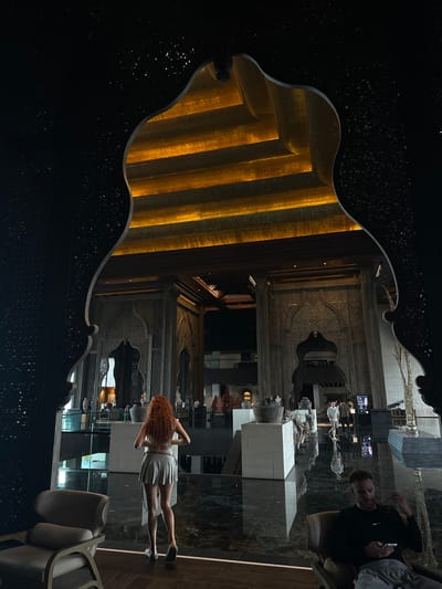 Captured in Nusa Dua, Indonesia, the image presents a reflected scene within a decorative, dark-toned frame. Inside the frame, illuminated vertical stripes of warm light contrast with the dark interior. The reflection encompasses a modern interior, including a woman in a light-colored dress and a man seated with a mobile phone. Art installations and a contemporary design are visible in the mirrored space. The environment suggests a luxury hotel or upscale resort lobby.