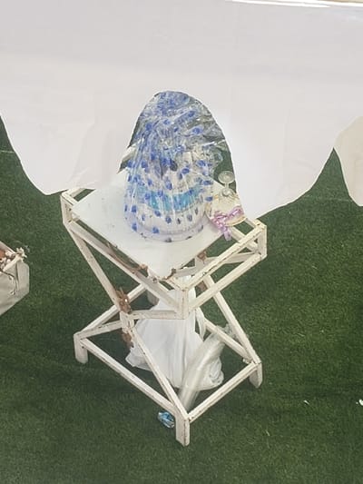 The scene depicts an outdoor event setting in Ajaokuta, Nigeria. A distressed white metal stand, exhibiting areas of rust and chipped paint, occupies the center-right of the frame. On its rectangular top surface rests a multi-tiered object, possibly a cake or a decorative centerpiece, encased in clear plastic wrap. This object is adorned with numerous small blue and white elements. Adjacent to it on the stand is a small, clear glass bell jar tied with a purple ribbon. Beneath the stand, on the green artificial turf, a white plastic bag and several clear plastic items, possibly discarded cups or bottles, are visible. To the far left, a portion of a white container is partially obscured. The upper portion of the image is dominated by a large white fabric sheet, draped to form a canopy or backdrop over the scene. No individuals are present in the observable area.
