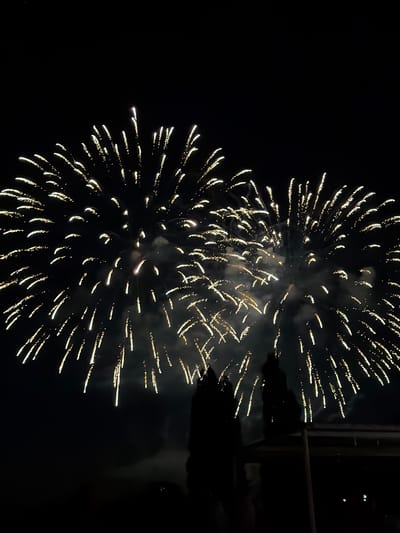 The image depicts a nighttime scene with two large, bright yellow fireworks exploding in a dark sky. The fireworks are detailed with numerous streaks of light radiating outwards, suggesting a vibrant display. In the foreground, silhouetted against the fireworks, are two figures. They appear to be standing, possibly on a rooftop or elevated platform, observing the spectacle. The overall atmosphere is one of celebration or a special event. The scene is set in the city of Brno, Czechia.