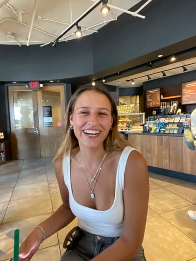 A young adult female is depicted smiling broadly and looking directly at the camera from an indoor setting identified as a Starbucks coffee shop, consistent with locations found in Washington, United States. She is light-skinned with medium-length light brown hair, wearing a white scoop-neck tank top, blue jeans, gold-colored hoop earrings, a silver bracelet on her left wrist, and two layered silver necklaces (one with a whale tail pendant, another with a light-colored rectangular pendant). Her mouth is open in a wide smile, revealing teeth, indicating an active state of laughter or cheerful conversation. A green straw from a beverage container is partially visible in the lower left corner, near her left hand. The background features typical Starbucks elements: a long wooden counter with coffee machines, product displays, and menu boards, a display case containing pastries, and a set of double wooden doors with glass panels marked with a red "EXIT" sign. The ceiling is dark gray with white exposed pipes and track lighting fixtures. A partial view of a lower leg and foot in a white sneaker is visible on the far right, indicating the presence of another individual.