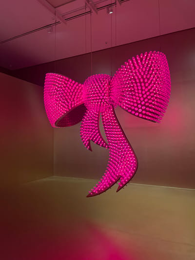 The image shows a large, bright pink bow sculpture hanging from the ceiling in a room. The sculpture is made up of numerous small, spherical lights that emit a vibrant pink glow. The room appears to be an art gallery or exhibition space, with neutral-colored walls and a ceiling with visible track lighting. The floor is also a neutral color and reflects the pink light from the sculpture. There are no people visible in the image. The sculpture is the dominant subject, casting a strong pink light that illuminates the surrounding space.

This artwork is located in the city of Málaga, Spain.