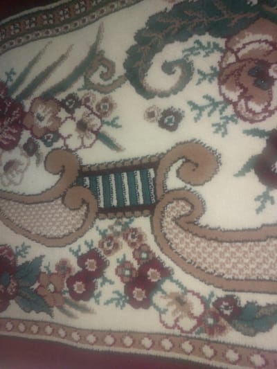 This is a close-up, high-angle, slightly blurry photograph of a portion of an ornate rug. The rug has a cream-colored background with a repeating pattern of stylized floral elements and scrolling motifs in shades of burgundy, green, and tan. The rug appears to be lying flat on a surface, possibly a floor. There are no people or other discernible objects visible in the image. The image provides no information about the location in the city of Ması, Kyrgyzstan, or any contextual details beyond the rug itself.