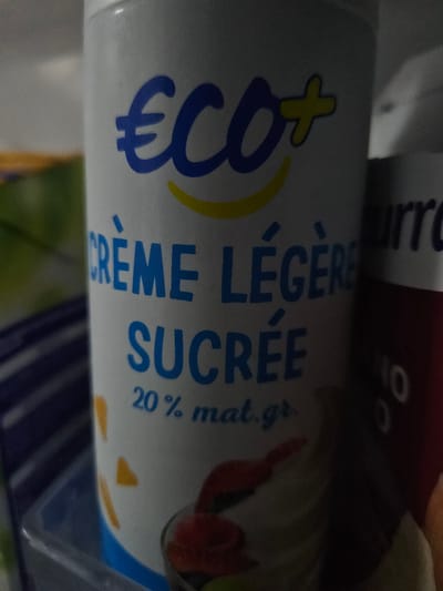 An extreme close-up, slightly tilted shot of the label on a white cylindrical container of what appears to be whipped cream. The logo at the top features a large blue "€" symbol followed by "CO+" in blue with a yellow outline. Below this, in blue text, are the words "CRÈME LÉGÈRE" followed by "SUCRÉE". Underneath this, the text "20 % mat. gr." is visible. The background is dark and out of focus, with hints of other containers or packaging. The image is dimly lit, with some glare on the container. The scene is not identifiable as being in Versailles, France, as no landmarks or contextual clues are present.