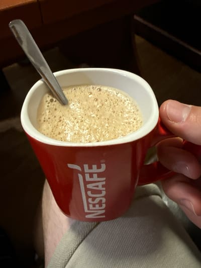 A close-up, high-angle shot captures a person's right hand holding a red mug filled with a foamy, light brown beverage, likely coffee. The mug has a white interior, and a silver spoon rests within the drink. The surface of the beverage is covered in a thick layer of small bubbles, indicating foam. The brand name "NESCAFÉ" is visible in white, uppercase letters on the red exterior of the mug, oriented upside down relative to the handle being held.
The hand, with visible fingers and a thumb, grips the handle of the mug. In the background, to the lower right, a textured, light-colored fabric, possibly clothing or upholstery in shades of light grey or beige, is partially visible. The upper left and lower right backgrounds are dark and out of focus, suggesting an indoor setting with subdued lighting, possibly a table or another surface. The image focuses on the act of holding the mug, possibly preparing to drink or after stirring. The scene is set in Hadzhidimitrovo, Bulgaria.