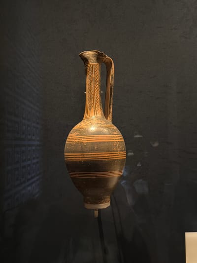 An ancient Greek terracotta amphora, characterized by its bulbous body, narrow neck, and handle, is displayed against a dark background. The amphora features black and orange geometric patterns, including horizontal bands on the body and a vertical, repeating motif on the neck. A faint, patterned reflection is visible on the left side of the dark background. The object is suspended by a thin rod, suggesting it is part of a museum exhibit. The overall lighting is dim, with a spotlight focused on the amphora. The specific location in Istanbul, Turkey, is not discernible from the image.