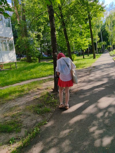 A young woman with bright pink hair is the sole subject of this image. She is seen from behind, wearing a loose white shirt over a bright red skirt. She has a canvas bag slung over her right shoulder and is wearing light brown sandals. The woman is leaning forward, appearing to inspect or tie something to the trunk of a large tree that stands to her left.
The setting is an urban environment with abundant greenery, suggesting a park or a tree-lined street. A light-colored building with numerous windows is visible on the left side of the frame, behind a patch of well-maintained grass. A paved walkway stretches into the distance on the right, bordered by trees. The ground in front of the trees is a mixture of dirt and grass. Sunlight filters through the dense tree canopy, casting dappled shadows across the ground and walkway, indicating it is daytime, likely a sunny afternoon. The overall mood is peaceful and ordinary, with no obvious signs of distress. There is no visible text in the image. The location context is Bucha, Ukraine.