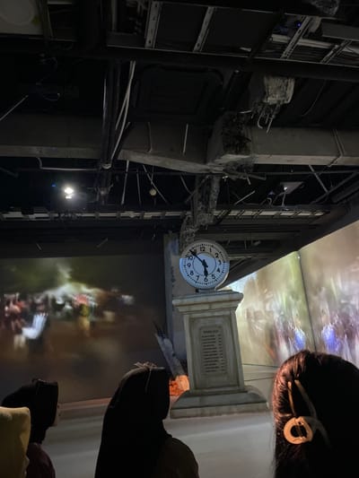 The scene is an indoor space with a large clock mounted on a tall column. The clock is black and white with Roman numerals and black hands. It is set at 2:50. The column is made of a light gray material, and has a plaque on the front side. The wall behind the column has a large projected image depicting a blurry, brightly colored scene of a city, possibly during a natural disaster. The image is not completely visible due to the position of the column, but it appears to be a city scene with buildings and people. The floor in front of the clock is white and smooth. There are three people in the scene, all are facing the clock and are obscured by darkness.  The three individuals appear to be standing on a white floor, observing the clock and the projected image. They are standing in the foreground of the image. The scene is lit by dim lights, casting long shadows from the ceiling. The ceiling has exposed beams and wires, and a portion of the ceiling appears damaged. The scene is located in an indoor space, possibly an exhibition hall or a museum. The location of the scene is Banguntapan, Indonesia, located at coordinates (latitude: -7.8, longitude: 110.407). The image was taken from a low angle, looking up at the clock.