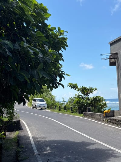 The image, taken in Kutuh, Indonesia, shows a curved asphalt road with a white dividing line. A white car is visible on the road. Lush green vegetation flanks the road, and in the distance, the ocean is visible beneath a blue sky with scattered clouds. The upper frame includes a portion of a concrete structure. A yellow object hangs from a roadside barrier.