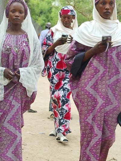 Three women wearing hijabs and long dresses are walking on a dirt road. The woman on the left wears a purple dress with white lace on top. The middle woman wears a dress with a white head covering and a dress with red and black flowers. The third woman is wearing a purple dress with white head covering and is holding a cell phone. All three women are looking down. A man is walking behind them. The location is Gandu Albasa, Nigeria.