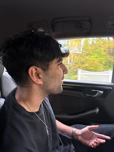 A male individual, approximately 20-35 years old, with dark, wavy hair and stubble, is seated in the rear passenger seat of a moving vehicle, looking out the window to the right. He wears a dark t-shirt, a silver-colored chain necklace with a cross pendant, and a silver-colored watch on his left wrist. His left hand is partially raised, with fingers extended. The vehicle interior features dark upholstery on the seat and door panel, with a grab handle visible on the ceiling. Outside the window, blurred autumnal foliage in green and yellow-orange hues is visible, along with a section of a white fence or railing. The scene is located within the city of Needham, United States.