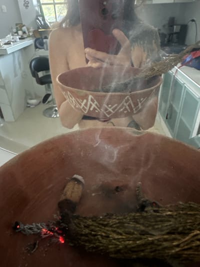 The image is a reflection in a mirror of a person holding a red heart-shaped object and a ceramic bowl with burning incense sticks inside. The bowl has white geometric patterns around its rim. The person's upper body, shoulders, and arms are visible. The background shows a domestic setting with a kitchen counter, cabinets, and a window. The specific location is stated to be in the city of Buenos Aires, Peru, though this is a geographical contradiction as Buenos Aires is in Argentina. The scene appears to be indoors, possibly a kitchen or dining area, with natural light coming from a window. There are no other people visible in the reflection or the environment. The focus is on the act of burning incense.