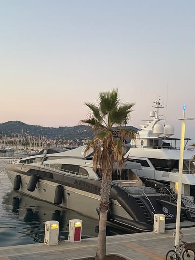 This media file captures a serene scene at a marina in Imperia, Italy, likely during the late afternoon or early evening. The primary subjects are large luxury yachts docked in the calm water, with a prominent palm tree in the foreground partially obscuring the view. The setting appears to be a waterfront promenade with a paved walkway.
The image showcases several large yachts, one of which is particularly detailed, showing its sleek, modern design with dark hull accents and white superstructure. Attached to its side is a large, black inflatable tender or dinghy. The yachts are moored alongside a concrete quay, where two white electrical hook-up boxes with illuminated lights are visible. One box has a red door, while the other has a white door.
In the background, a hillside town with numerous buildings and sailboats dotting the harbor creates a picturesque backdrop. The sky is a soft gradient of pale blue and warm, peachy tones, suggesting the approach of sunset. The weather appears calm and clear. A bicycle is leaning against the railing of the promenade, hinting at the presence of recreational activity.
There is visible text on the side of the large inflatable tender, which appears to be "DIANA VALLETTA" in white lettering. The overall mood is tranquil and peaceful, characteristic of a quiet harbor at dusk.